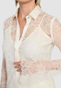 Blonde bluse med lange gennemsigtige ærmer, elfenbensfarvet bund, knaplukning fortil, blomster mønster, og en satin krave med dekorative knapdetaljer.