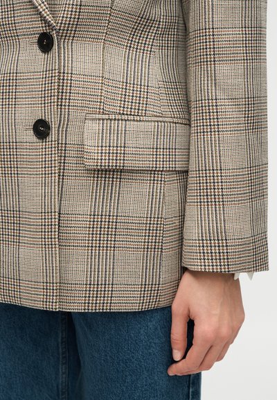 Nahaufnahme eines beigen und braunen karrierten Blazers mit schwarzen Knöpfen und einer Klappentasche, getragen über blauen Jeans.