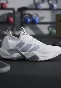Scarpa da corsa Adidas bianca e grigia su un pavimento da palestra con kettlebell colorati sfocati sullo sfondo.