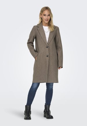 Manteau en mélange de laine marron avec un col à revers, deux poches avant et deux boutons noirs. Porté par-dessus un haut blanc et associé à un jean foncé et des bottes noires.