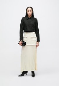 Žena s pletenými vlasmi v krátkom čiernom dvojradovom saku, krémovej plisovanej maxi sukni s prednými vreckami, čiernych špicatých čižmách, držiaca čiernu listovú kabelku.