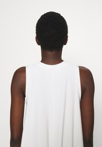 Personne aux cheveux courts et bouclés portant un haut blanc sans manches, vue de dos sur un fond clair uni.