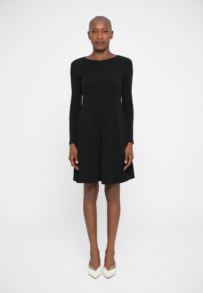 Robe noire à manches longues au design ajusté, avec un col rond et une jupe évasée. Associée à des escarpins blancs à bout pointu.