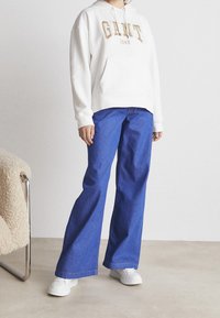 Sudadera blanca con letras doradas, combinada con jeans de pierna ancha azules. El modelo lleva zapatillas blancas, de pie sobre un suelo neutro con muebles suaves cercanos.