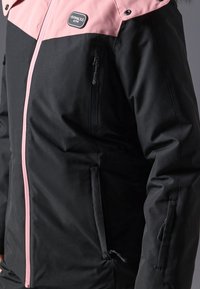 Svart og rosa jakke med glidelås, med en kontrasterende rosa øverste del, flere lommer og en glatt tekstur. Har en merkevarelogo-lapp.