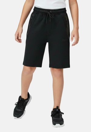 Kind draagt zwarte knielange shorts met ritssluitingzakken, zwarte sneakers en een wit topje, staand tegen een effen achtergrond.
