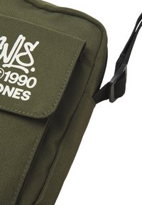 Bolsa de mensajero de nylon verde oliva con solapa, logo blanco, correa ajustable y superficie texturizada. Presenta un broche lateral para sujeción.