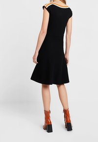 Žena v černých šatech ve tvaru A s oranžovým a bílým lemováním na ramenech, spárovaná s lesklými hnědými kotníkovými botami s blokovým podpatkem.