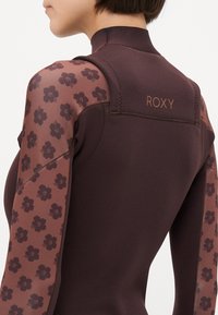Costum de neopren maron cu mâneci transparente cu model floral și corp texturat. Are brandul Roxy pe spate, subliniind designul și contrastul de culoare.