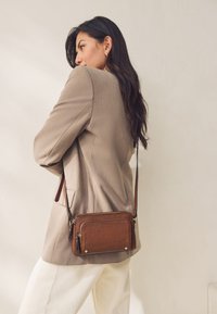Sac bandoulière en cuir marron avec une finition texturée, deux compartiments zippés, des accents dorés et une sangle d'épaule amovible.