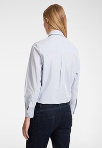 Camicia di cotone azzurro chiaro con pieghe sul retro, maniche lunghe e colletto classico. Abbinata a jeans scuri, che mostrano un design semplice.