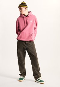 Haut à capuche rose avec poche avant et logo blanc, associé à un pantalon large marron foncé et des baskets vertes/blanches. Bonnet rayé sur la tête.
