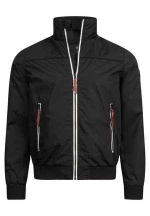Homme portant une veste bomber noire sur un t-shirt blanc, un jean bleu foncé et des baskets blanches, debout avec une main dans la poche.