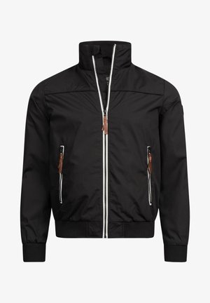 Homme portant une veste bomber noire sur un t-shirt blanc, un jean bleu foncé et des baskets blanches, debout avec une main dans la poche.