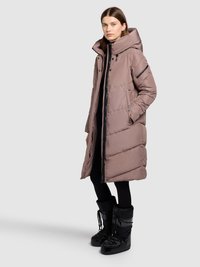 Cappotto lungo imbottito rosa con collo alto e cerniera frontale; presenta tasche laterali e dettagli neri a contrasto; indossato sopra pantaloni e stivali neri.