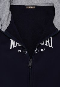 Giacca zip-up blu navy con cappuccio grigio, realizzata in materiale morbido. Presenta un logo e numeri bianchi sul davanti e una cerniera liscia.