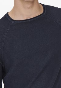 Gros plan sur une personne portant un haut à manches longues bleu marine texturé avec un col rond, montrant le cou et le haut de la poitrine.