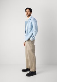 Light blue denim jacket with two chest pockets, paired with beige wide-leg pants and black lace-up shoes, against a plain light background.