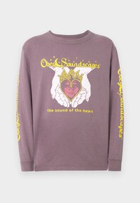 Ilgomis rankovėmis purpurinė medvilninė marškinėliai, su grafiniais atvaizdais, kur rankos laiko širdį su liepsnomis, ir geltonu tekstu priekyje ir ant rankovės.