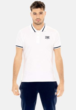 Giovane uomo che indossa una polo bianca con bordino nero e logo, abbinata a pantaloni blu scuro su uno sfondo uniforme.