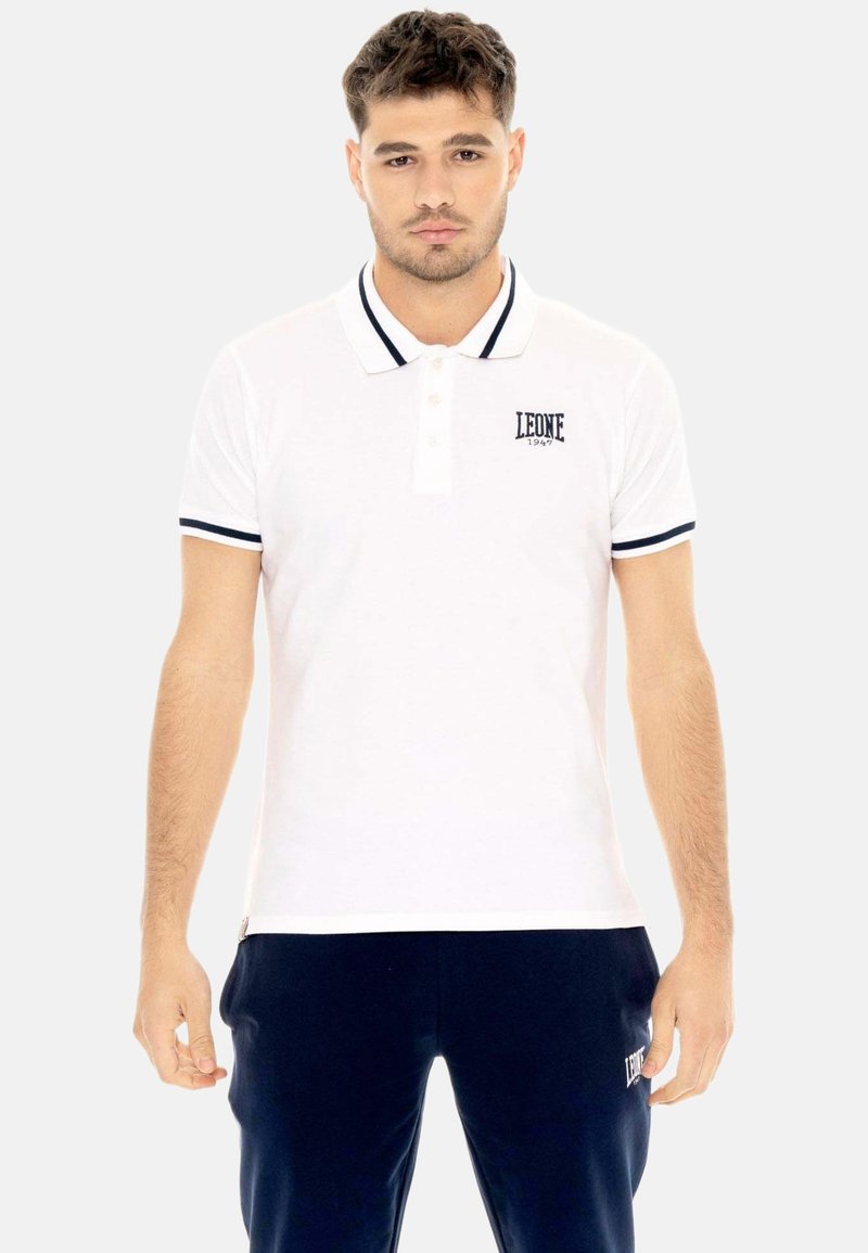 Jeune homme portant un polo blanc avec bordure noire et logo, assorti à un pantalon bleu marine foncé sur un fond uni.
