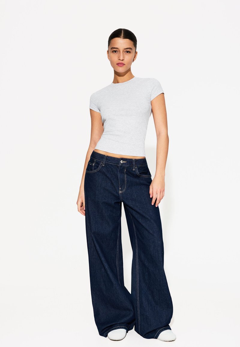 Tricou ajustat de culoare gri deschis combinat cu blugi evazați de culoare albastru închis. Blugii au buzunare frontale și cusături în contrast.