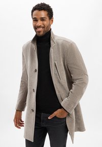 Uśmiechnięty mężczyzna w beżowym zapinanym płaszczu na czarnym swetrze polo i ciemnych dżinsach, trzymający jedną rękę w kieszeni.