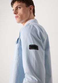 Hele sinine jakk, millel on lõdva istuvusega lõige, vasakul varrukal musta kangast logopatch ja ees klapptasku.