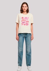 Cream short-sleeve t-shirt with graphic design featuring pink hearts, flowers, and text. Paired with light blue loose-fitting jeans and white sneakers.