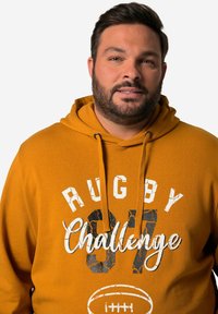 Orange hoodie med snoretræk i hætten, med grafisk tekst "RUGBY 97 Challenge" i kontrasterende sort og hvid, med et fodboldikon nedenfor.
