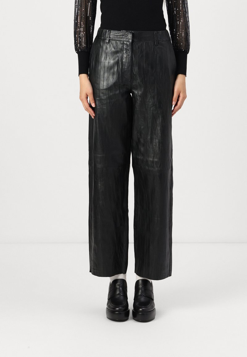 Sorte cropped bukser med vide ben lavet af skinnende tekstureret læder, med en plisseret design og en tætsiddende talje. Sammen med kraftige sorte sko.