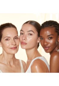 Three women with diverse skin tones and medium-length hair, wearing light-colored, sleeveless tops, smiling with fresh, natural-looking skin.