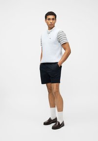 Moški, oblečen v belo telovnik z vzorcem kit, čez tigrasto črtasto majico, mornarsko modre kratke hlače, bele nogavice in rjave loaferje, stoji na belem ozadju.