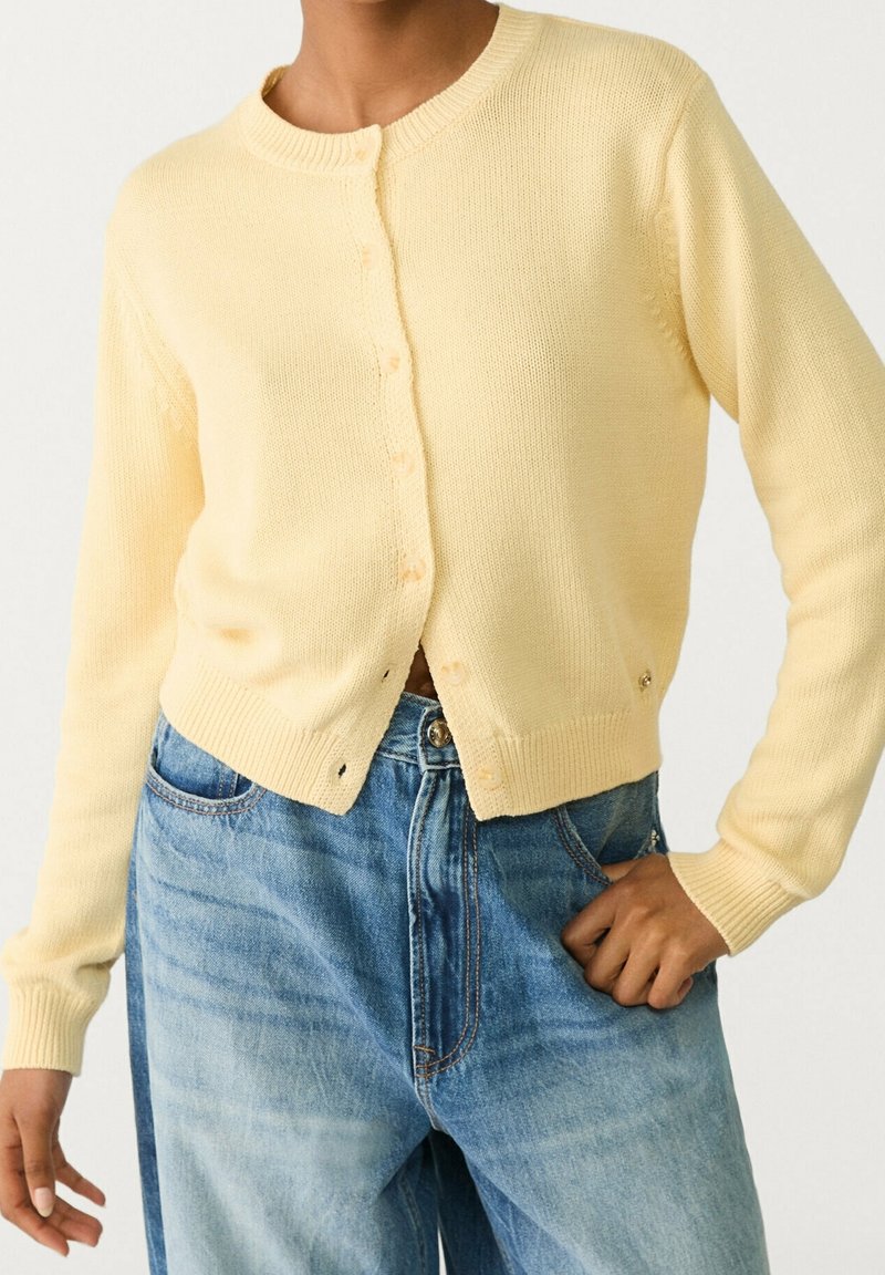 Personne portant un cardigan boutonné jaune pâle et un jean taille haute bleu clair, une main dans la poche et l'autre détendue.