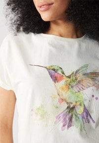 Sidentröja med en akvarellillustration av en kolibri i gröna, lila och gula nyanser, med en avslappnad halsringning.