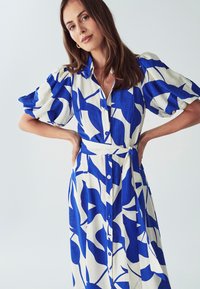 Mėlynos ir baltos spalvų raštuota suknelė su trumpomis pūstomis rankovėmis, mygtukų užsegimu, surišamu juosmens diržu ir srauni siluetu, pagaminta iš tekstūruoto audinio.