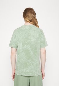 Osoba z długimi włosami, nosząca jasnozieloną koszulę z krótkim rękawem w stylu tie-dye oraz pasujące zielone spodnie, stojąca przodem do białego tła.