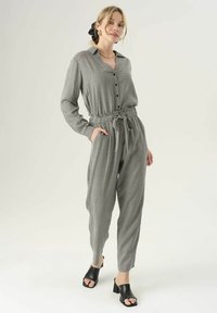 Grijze jumpsuit met een patroon, kraag, knopen aan de voorkant, elastische taille en zijzakken. Gedeeld met zwarte sandalen met blokhakken.