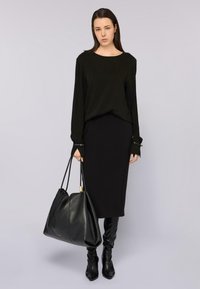 Donna con top nero a maniche lunghe, gonna midi nera e stivali neri fino al ginocchio, mentre tiene in mano una grande borsa di pelle nera.