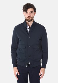 Giacca blu navy realizzata in tessuto morbido, con colletto alto, chiusura a bottoni e due grandi tasche frontali con pattine. Polsini e orlo texture.