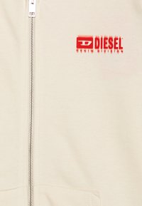 Giacca in cotone crema con zip, caratterizzata da una zip argentata e un logo Diesel rosso stampato sul petto. Texture liscia senza motivi.