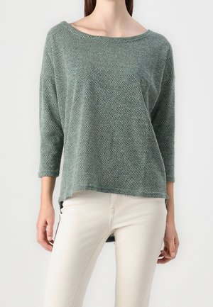 Femme portant un pull texturé vert sauge à manches longues avec un col bateau et un pantalon blanc ajusté sur fond uni.