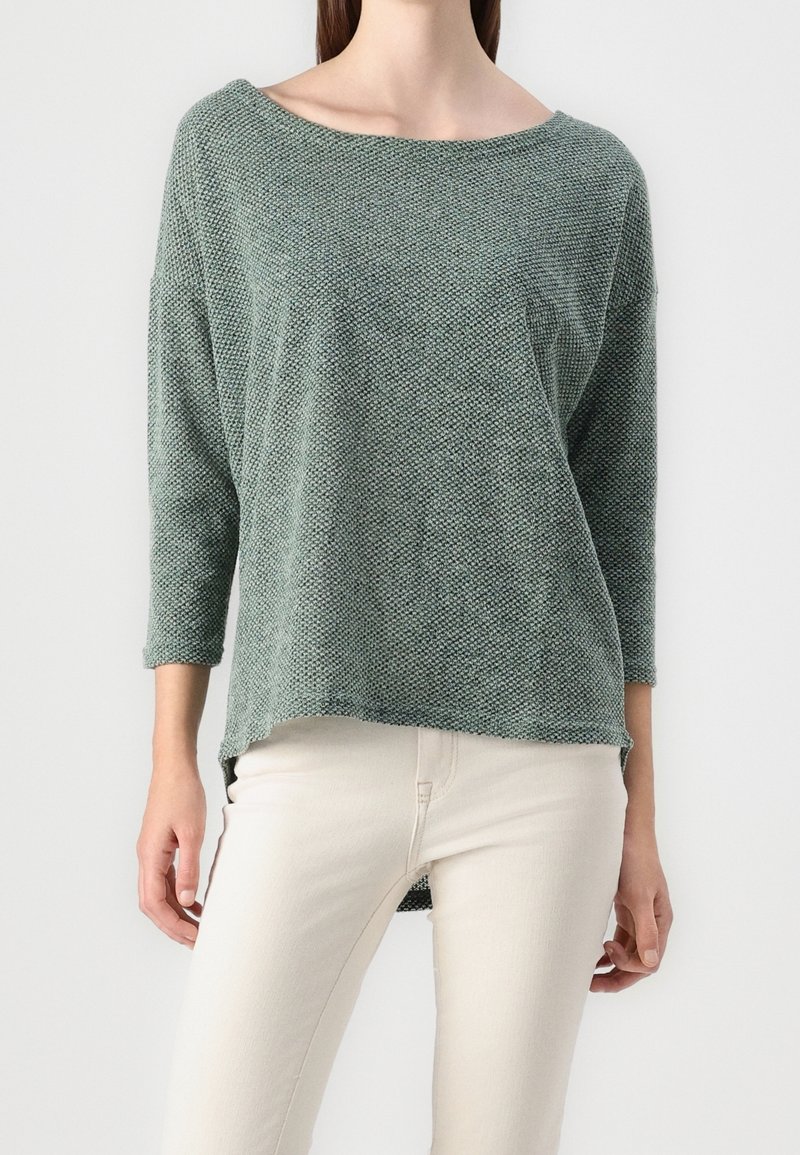 Femme portant un pull texturé vert sauge à manches longues avec un col bateau et un pantalon blanc ajusté sur fond uni.