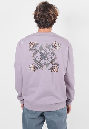 Personne aux cheveux courts et bruns porte un sweat-shirt violet clair avec un grand motif floral en nuances de gris et d’orange dans le dos.
