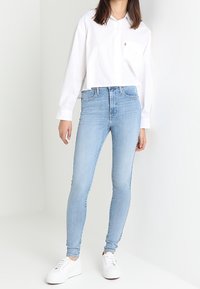 Mujer vestida con una camiseta blanca corta, jeans ajustados de color azul claro y zapatillas blancas, de pie contra un fondo blanco liso.