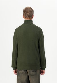 Mann mit kurzen, lockigen Haaren, der einen dunkelgrünen, gerippten Rollkragenpullover und dunkel gewaschene Jeans trägt, steht mit dem Rücken zu einem weißen Hintergrund.