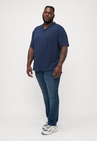 Camisa de manga corta azul marino con un ajuste relajado y cierre de botones, combinada con jeans azul oscuro y zapatillas deportivas grises.