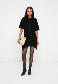 Μαύρο ριμπ κοντό φόρεμα δύο τεμαχίων με κοντά μανίκια, κλείσιμο με κουμπιά και ασύμμετρο στρίφωμα, συνδυασμένο με διάφανα μαύρα καλσόν και κίτρινη τσάντα.