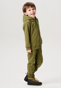 Olijfgroen hoodie met knopen aan de voorkant, gecombineerd met bijpassende tapered broek; gedragen met zwarte laarzen met gele veters en grijze accenten.