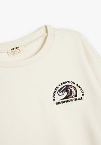 Sudadera de algodón crema con un diseño de ola bordado y texto: "LAS VACACIONES DE VERANO ESPERAN EN ESTE REFUGIO EN EL MAR."